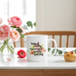 A white ceramic mug with the printed quote 'Behind every successful woman is herself' in black script, surrounded by a watercolor floral design. The mug is styled on a white table with pink roses, a pink rose, and a muffin in a kitchen setting.