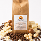 A kraft paper bag of Good Morning Coffee & Tea White Chocolate Macadamia Nut Coffee stands upright on a white background, surrounded by coffee beans, white chocolate chunks, and macadamia nuts.