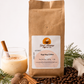 A kraft paper bag of Great Mornings Coffee & Tea labeled Egg Nog Coffee stands on a white surface with coffee beans, cinnamon sticks, a glass of egg nog, and evergreen branches around it.