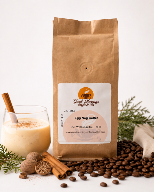 A kraft paper bag of Great Mornings Coffee & Tea labeled Egg Nog Coffee stands on a white surface with coffee beans, cinnamon sticks, a glass of egg nog, and evergreen branches around it.