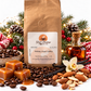Great Mornings Holiday Cheer Coffee in a kraft paper bag on a white studio background, surrounded by roasted coffee beans, caramel pieces, vanilla beans, toasted almonds, a small bottle of rum, evergreen branches, pinecones, and warm holiday lights.