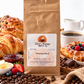 A brown kraft paper bag of Great Mornings Coffee & Tea Breakfast Blend stands centered on a white surface surrounded by coffee beans, pastries, a cup of coffee, jam, butter, and berries. The bag label reads Great Mornings Coffee & Tea and Breakfast Blend.