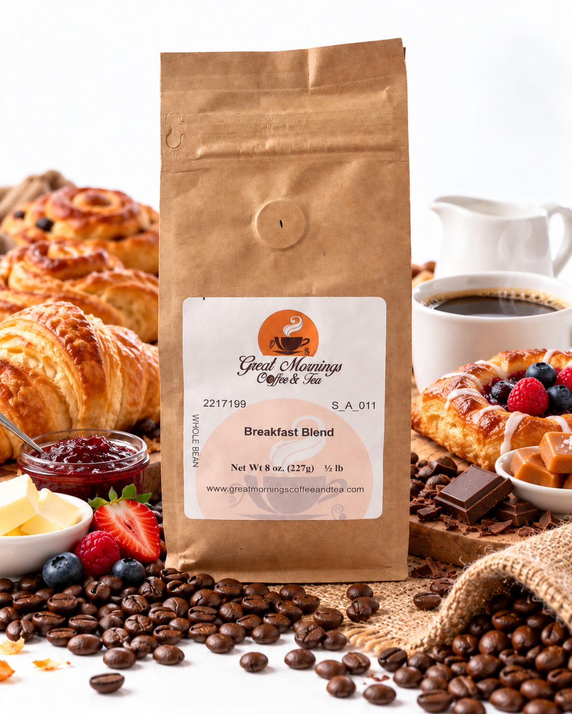 A brown kraft paper bag of Great Mornings Coffee & Tea Breakfast Blend stands centered on a white surface surrounded by coffee beans, pastries, a cup of coffee, jam, butter, and berries. The bag label reads Great Mornings Coffee & Tea and Breakfast Blend.