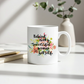A white ceramic mug with a colorful floral design and the quote 'Behind every successful woman is herself.' in black script, placed on a white surface with books and a vase in the background.