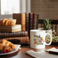 A white ceramic coffee mug with a colorful floral design and the text 'Behind every successful woman is herself', placed on a wooden table next to croissants, books, and a notebook.